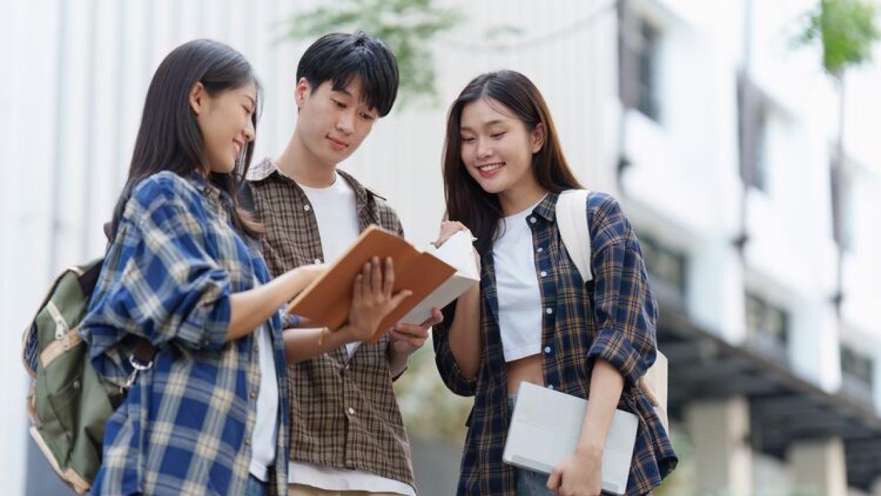 Three young people, two women and one man, standing outside looking at an open book.