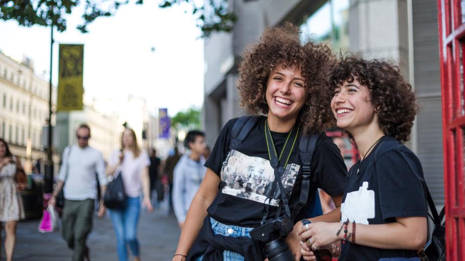 Two young women smiling, standing outside a red phone booth in London.