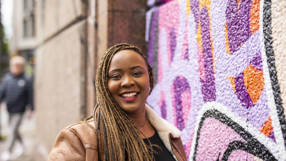 A young black woman standing against a colourful, graffiti-painted wall and smiling
