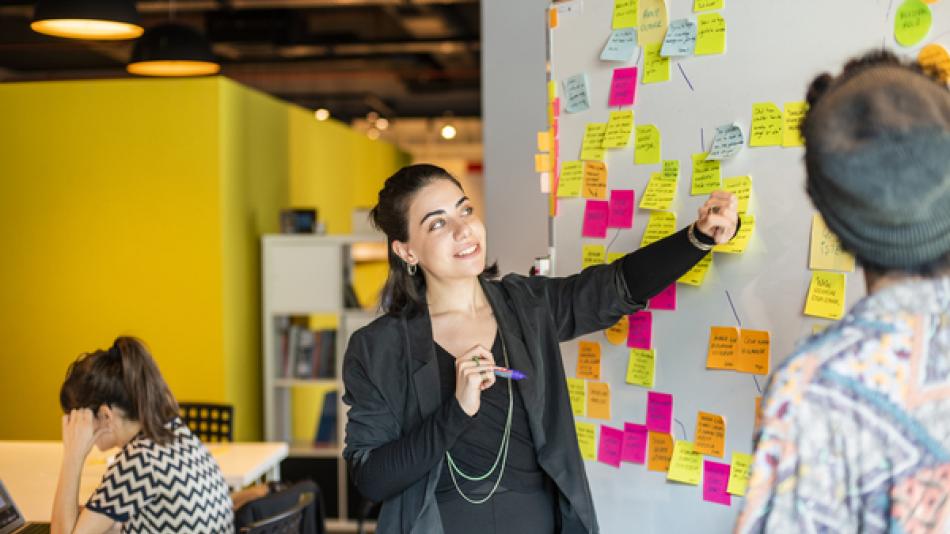 A young woman inside pointing to a whiteboard with colourful stick-on notes, explaining work processes to another male colleague in the office
