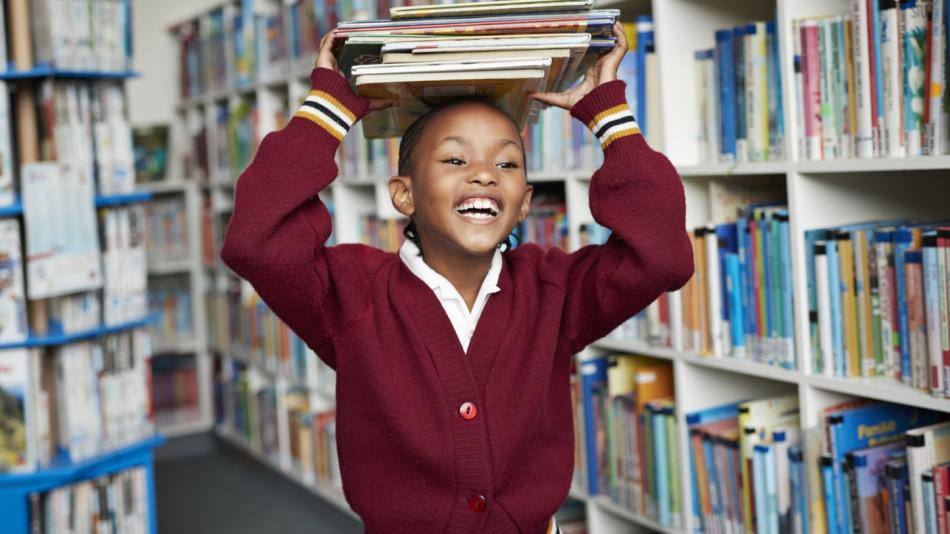 A smiling child wearing a red sweater in a library, holding books on top of her head