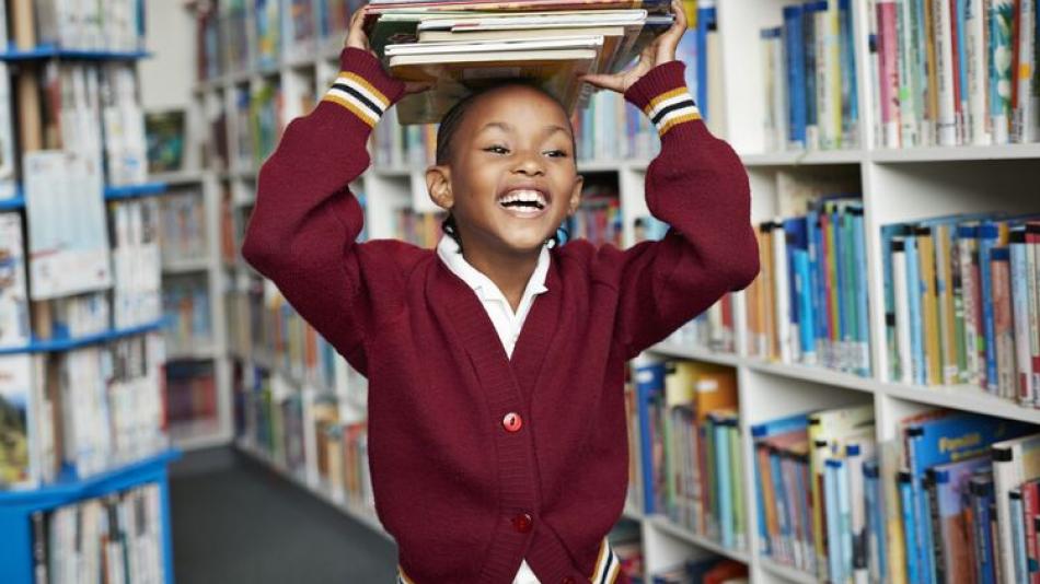 Photograph of a school girl smiling and balancing stack of books on her head in a library