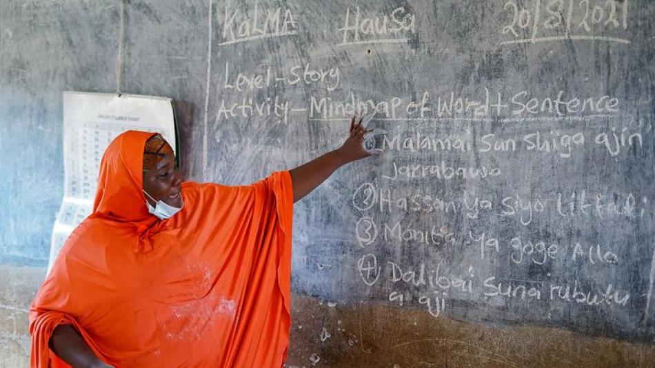 This image shows a teacher in an orange hijab standing in front of a chalkboard, pointing at some text written on it. The chalkboard contains both English and Hausa writing.  The text on the board includes:  Kalma Hausa 20/12/2021  Level: Story  Activity: Mindmap of Word The Kano Literacy and Math Accelerator (KaLMA) Britishcouncil.org https://www.britishcouncil.org › partner › track-record The Kano Literacy and Maths Accelerator (KaLMA) programme is dedicated to improving education in Kano State, Nigeria. 
