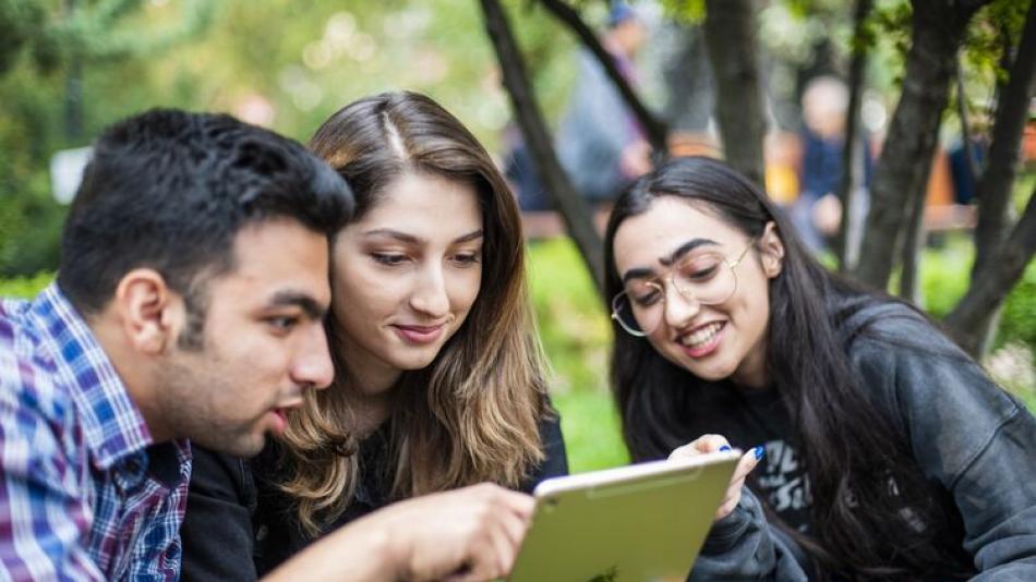 Three young adults in a park looking at an Ipad.